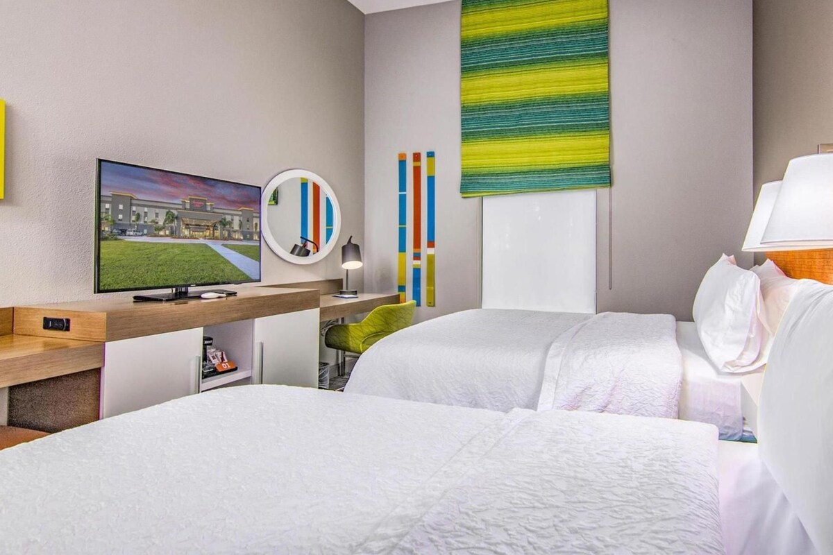 Habitación de hotel con dos camas, televisión, escritorio con computadora, espejo y vista a un edificio.