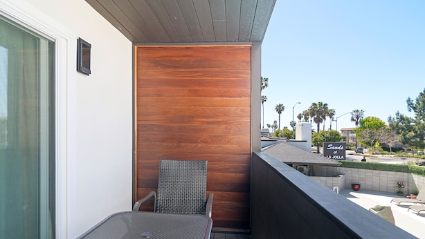 Un balcón con una mesa y silla de metal, un muro de paneles de madera y vista a un estacionamiento con un letrero que dice "Sands of Jolla".