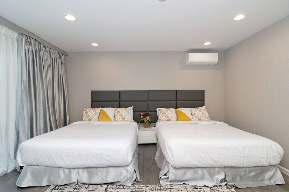 Ein Doppelzimmer mit zwei Betten, einem dunklen Kopfteil, einem kleinen Nachttisch mit Blumen und einer Klimaanlage an der Wand.