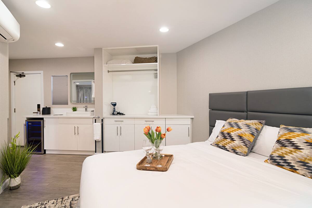 Ein modernes Hotelzimmer mit einem großen Bett, einem Nachttisch mit Tablett, auf dem zwei Weingläser und Blumen stehen, einem eingebauten Kleiderschrank und einer kleinen Küchenzeile.