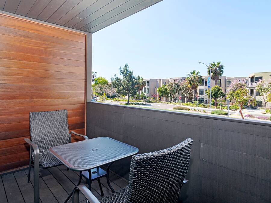 A balcony with two wicker chairs and a table, overlooking a residential area with trees and buildings.
