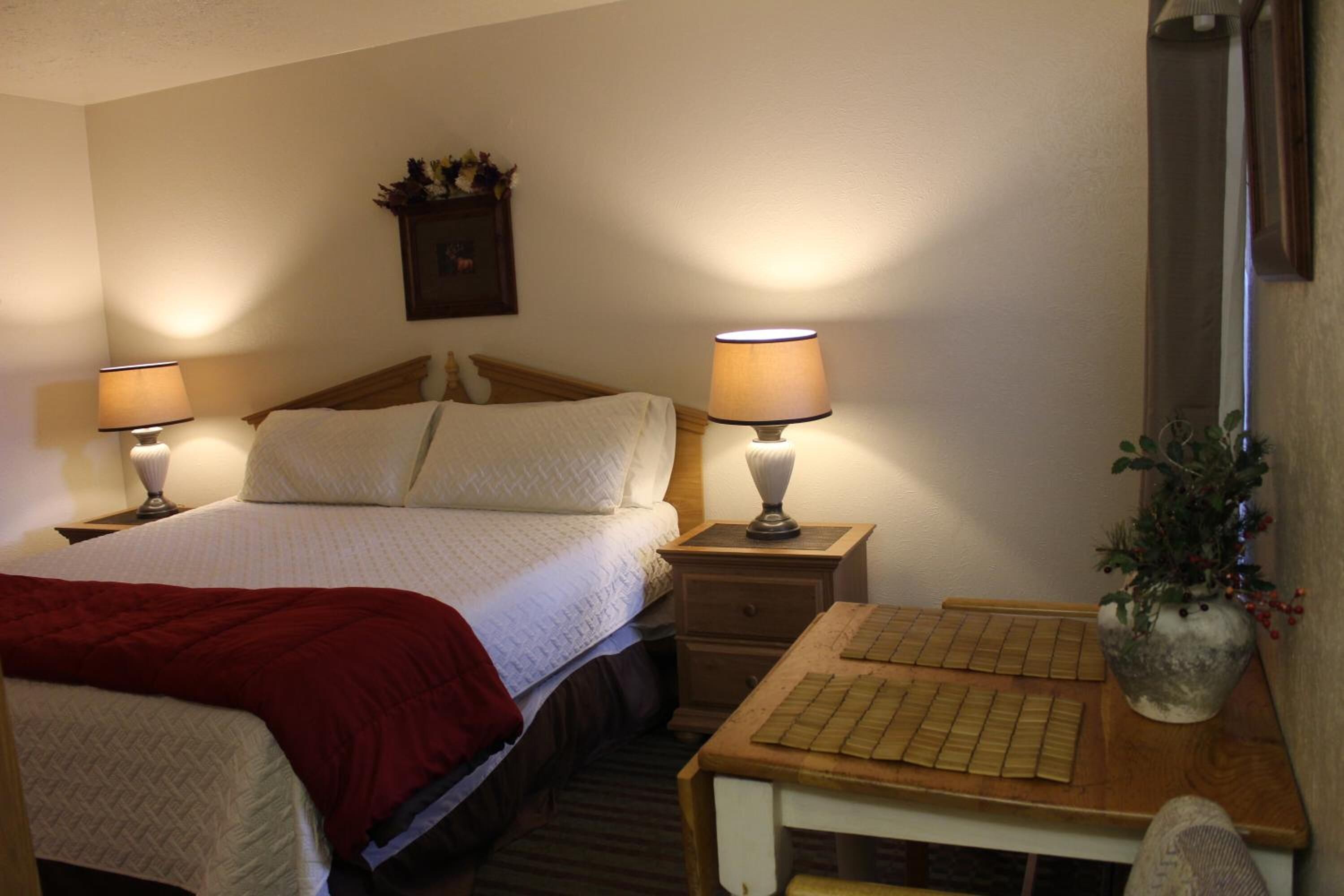 A hotel room with a bed, two bedside lamps, a wooden nightstand with a keyboard, a red throw blanket, and a potted plant.