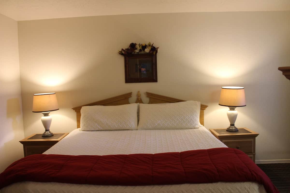 Een net opgemaakt bed met een houten hoofdbord, twee nachtlampjes en een aan de muur bevestigd fotolijstje.