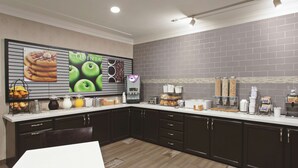 Cozinha com armários escuros, bancada branca e painel de azulejos. Há diversos alimentos e eletrodomésticos na bancada.