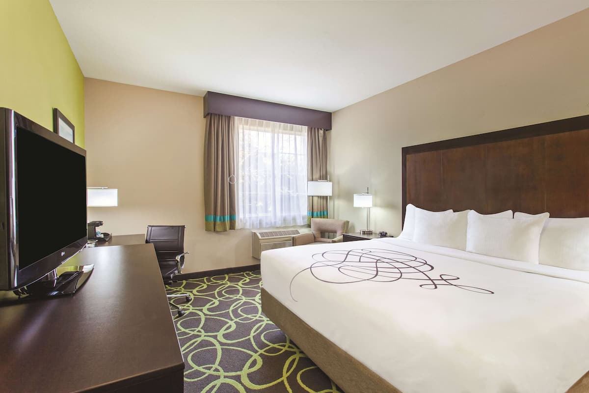 A hotel room with a large bed, a desk with a flat-screen TV, a chair, a desk lamp, and a window with curtains.