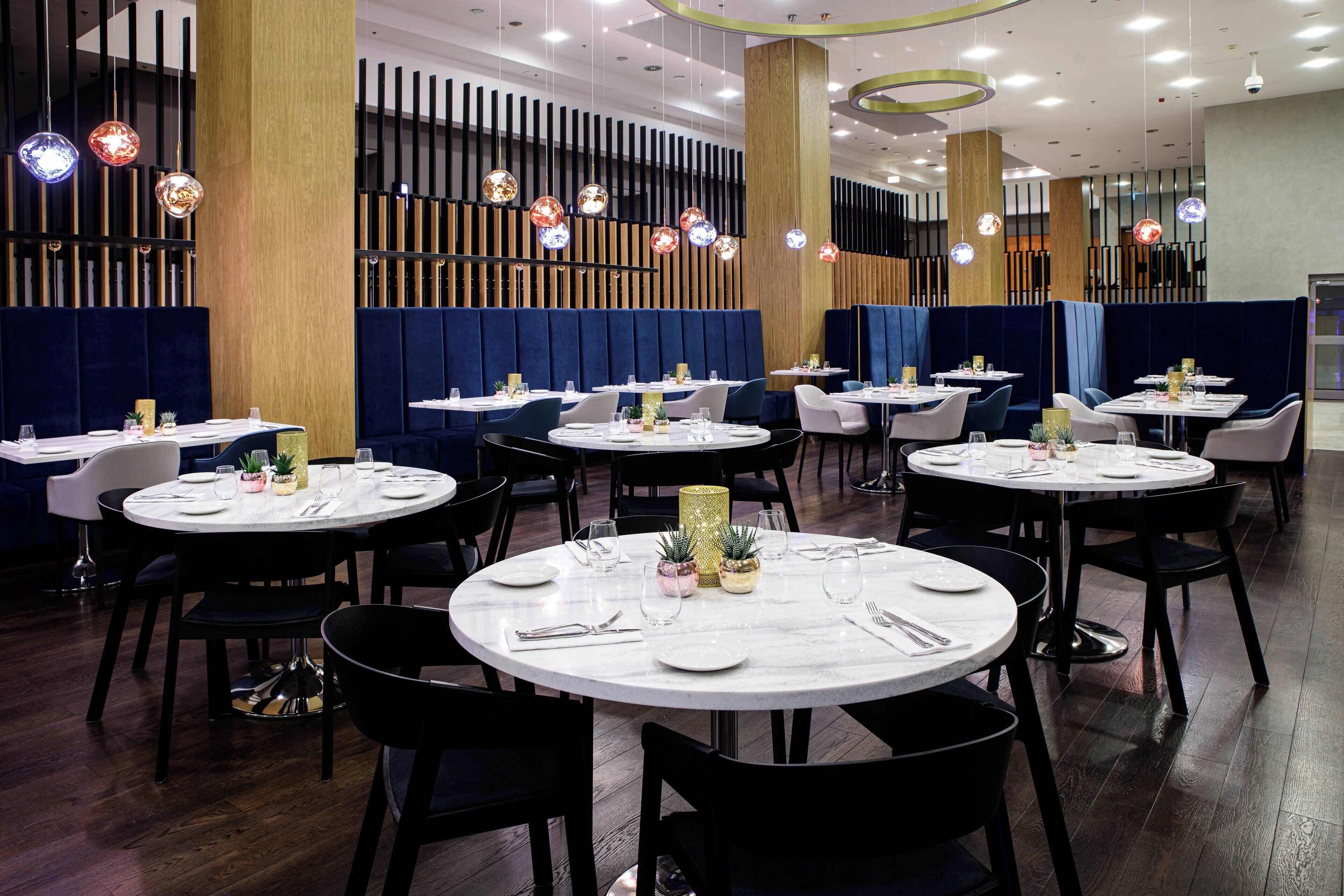 Un restaurante con mesas redondas, sillas negras y asientos tapizados en azul.
