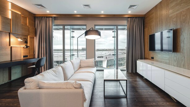 Un salon moderne avec un canapé, un téléviseur fixé au mur et une vue sur la ville grâce à de grandes fenêtres.