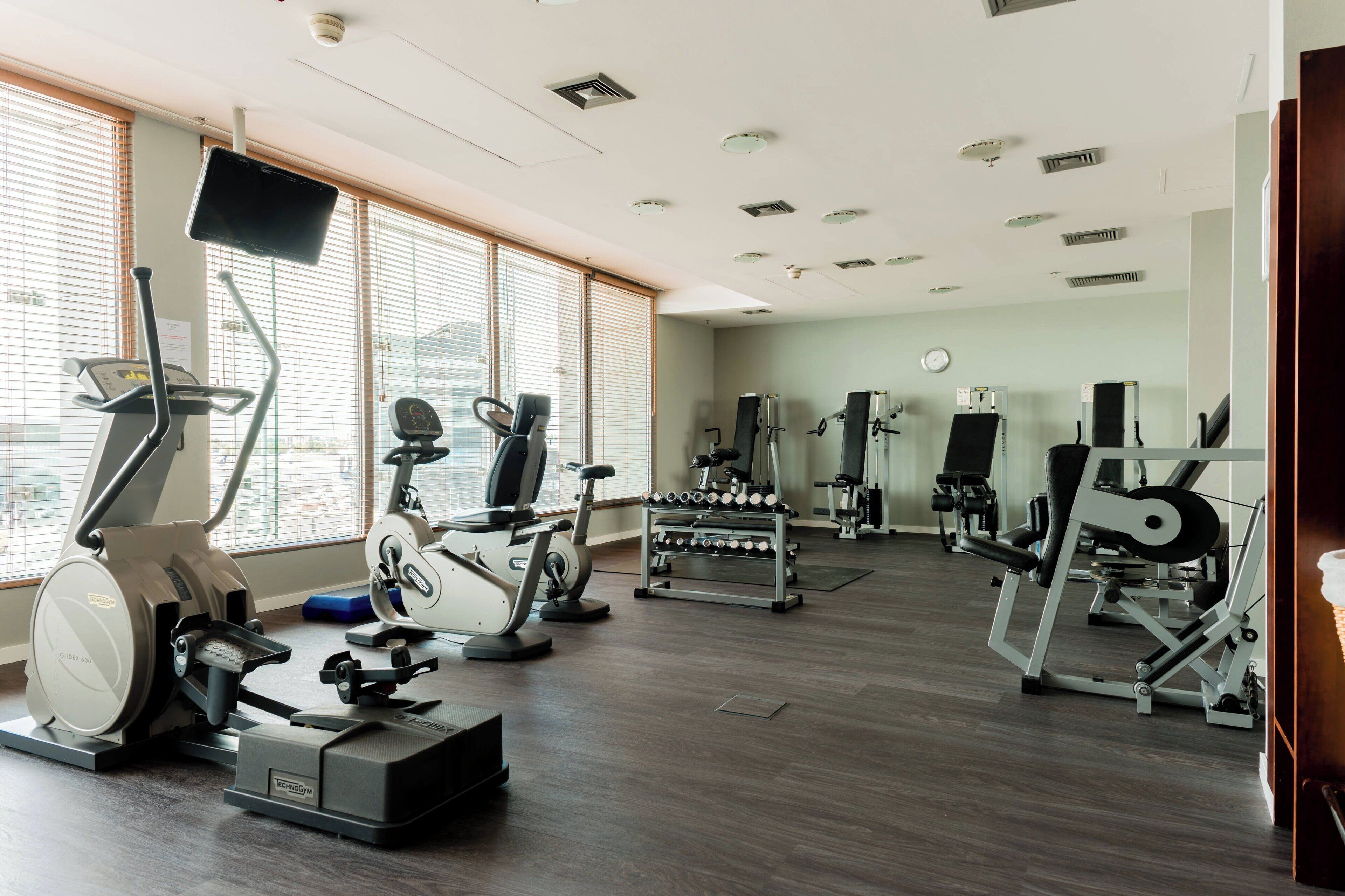 A gym with cardio machines, weight stations, and a large window with blinds.