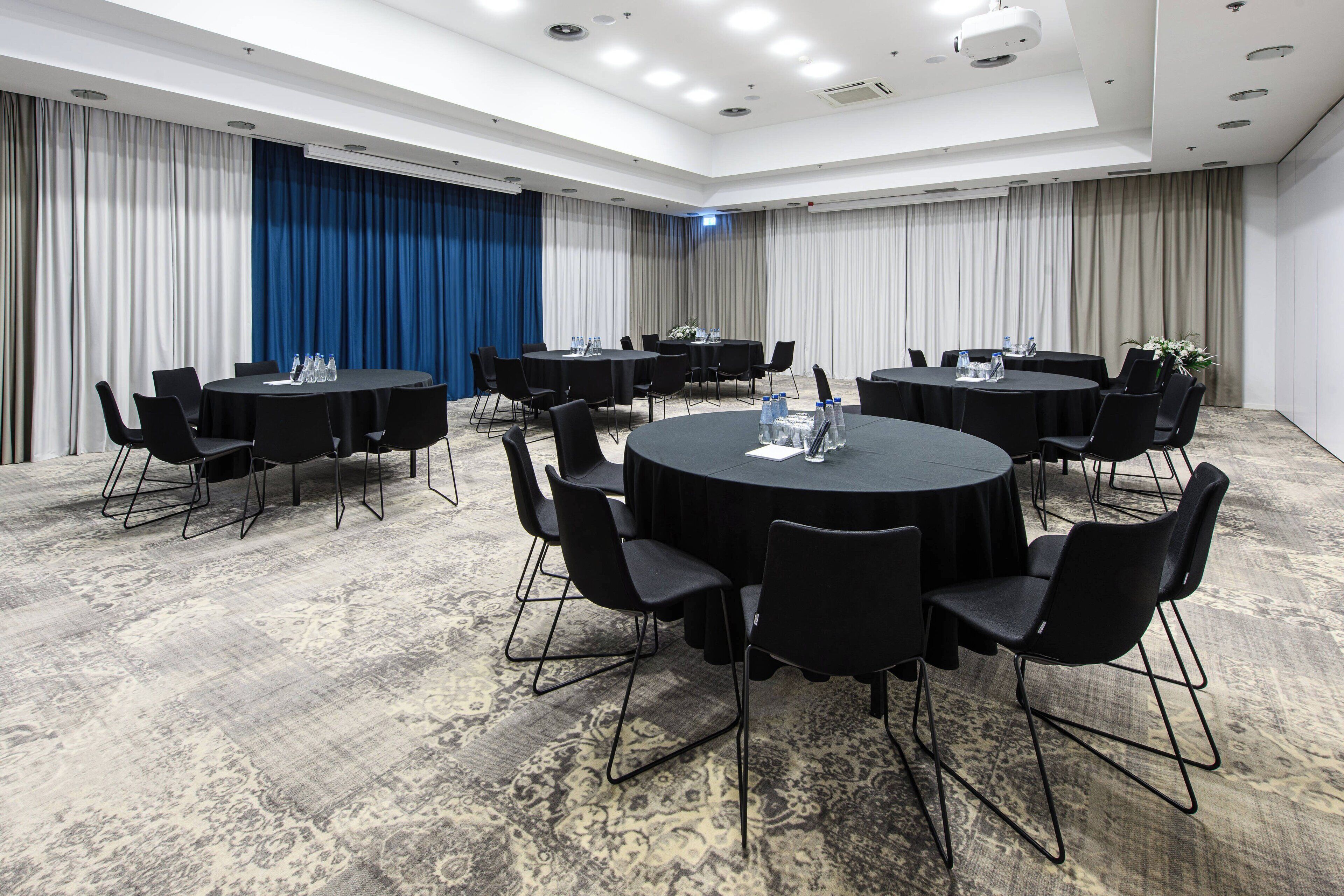 A conference room with round tables and black chairs, equipped with water bottles and a projector.