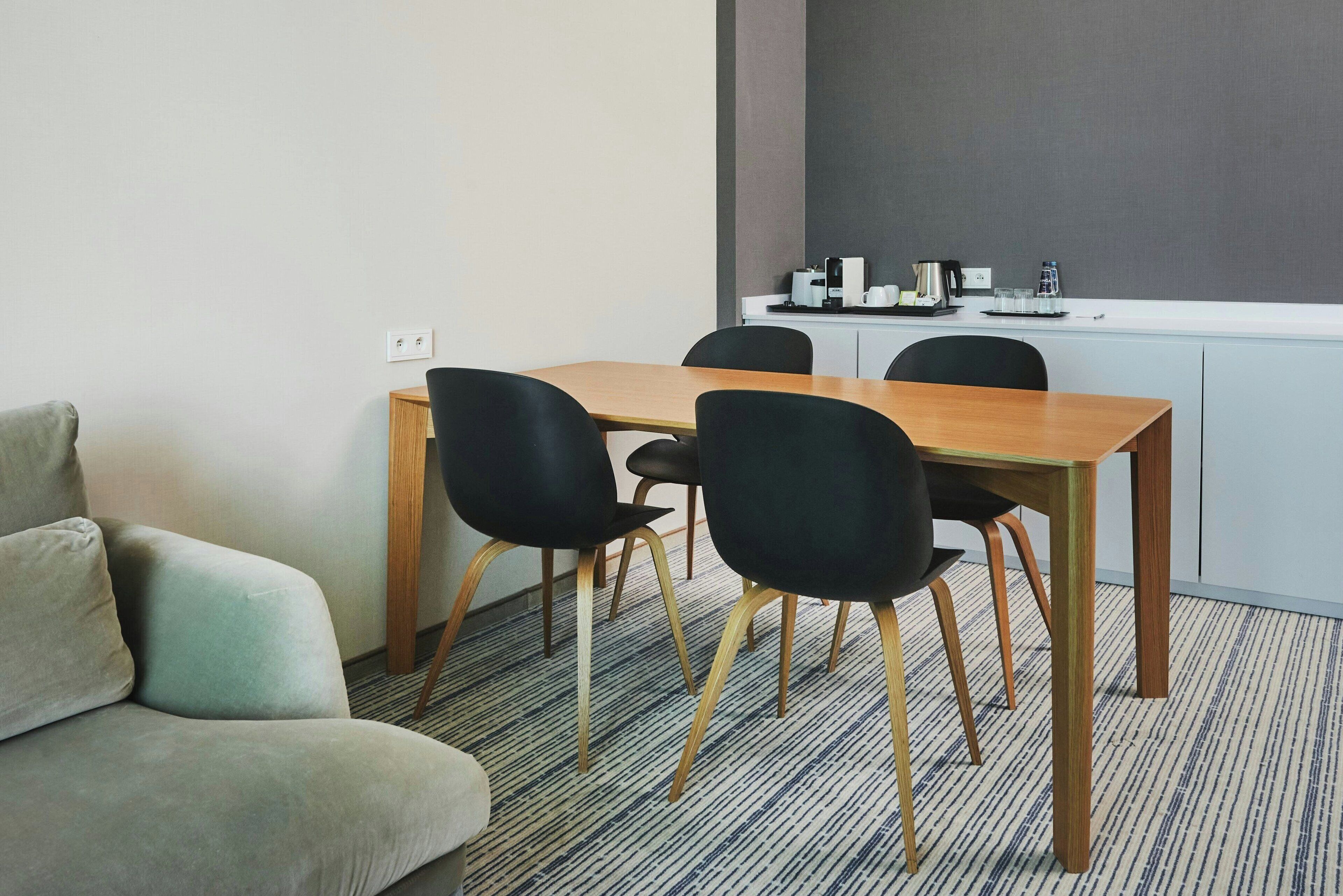 A modern room with a wooden table and four black chairs, a grey sofa, and a kitchenette with coffee and water dispensers.