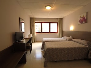 Habitación de hotel con dos camas, un escritorio con televisor, una ventana con cortinas y una lámpara fijada a la pared.