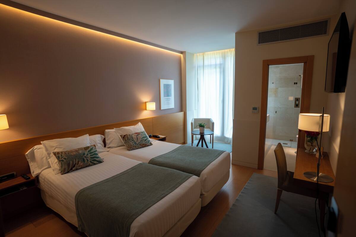 A hotel room with two beds, a desk, a chair, and a small table with a lamp.