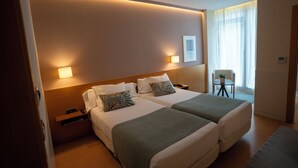 Una habitación de hotel con dos camas, mesitas de noche, una pequeña mesa con una planta y una ventana con cortinas.