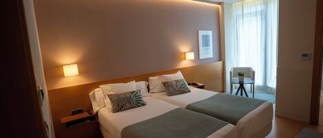 A hotel room with two beds, bedside tables, a small table with a plant, and a window with curtains.