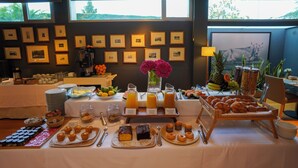 Un bufé de desayuno con diversas opciones de comida y bebidas, fotografías enmarcadas en la pared y vistas al exterior con vegetación.