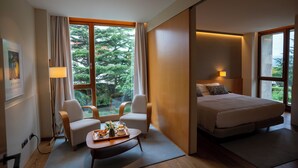A modern hotel room with a bed, two chairs, a small table with breakfast, and large windows offering a view of trees.