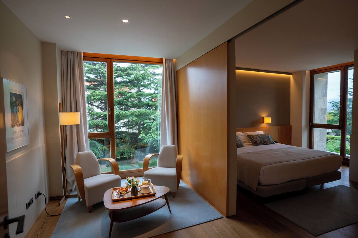 A modern hotel room with a bed, two chairs, a small table with breakfast, and large windows offering a view of trees.