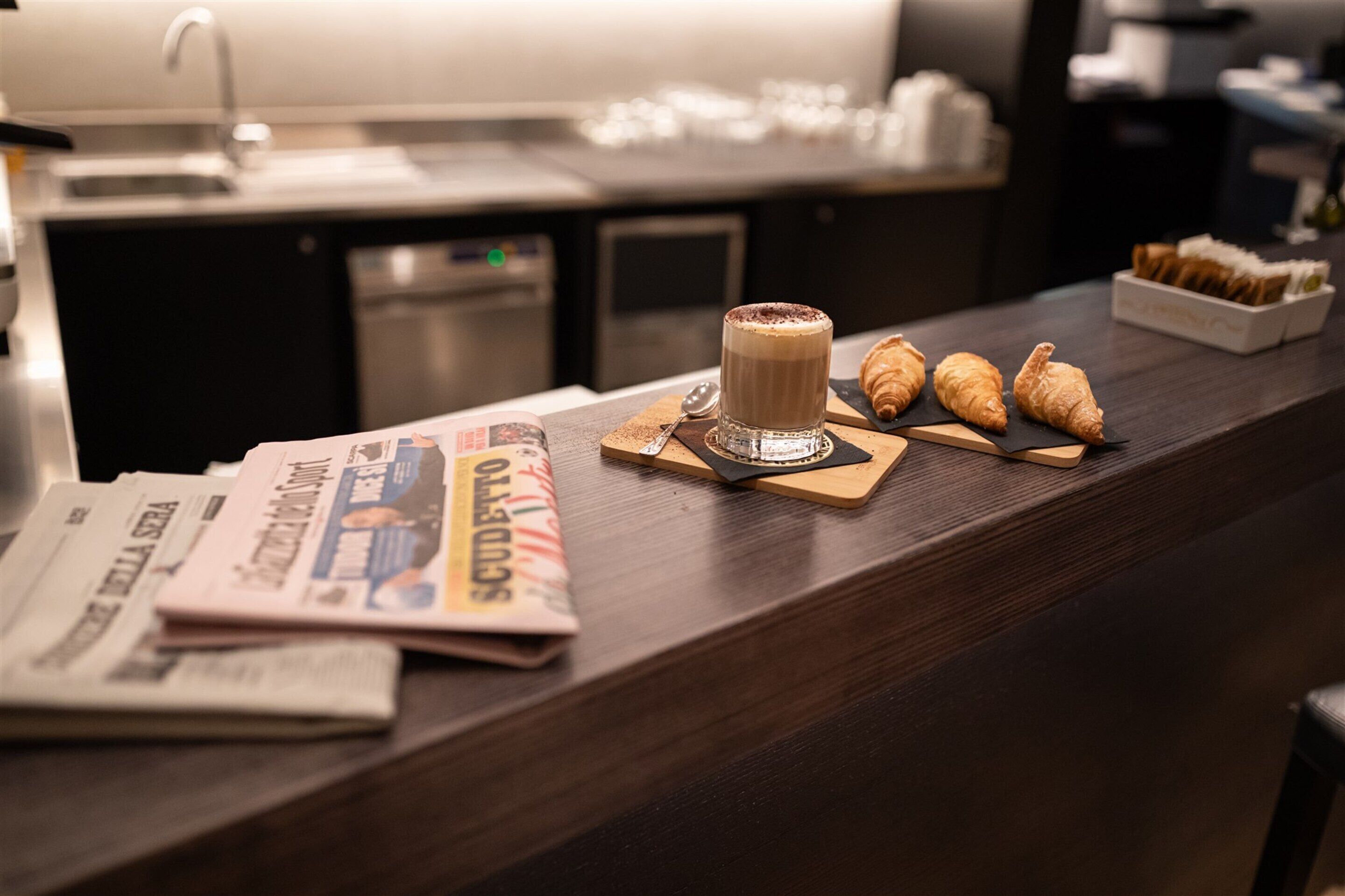 Un coin bar avec une tasse de café, des croissants et des journaux sur le comptoir.