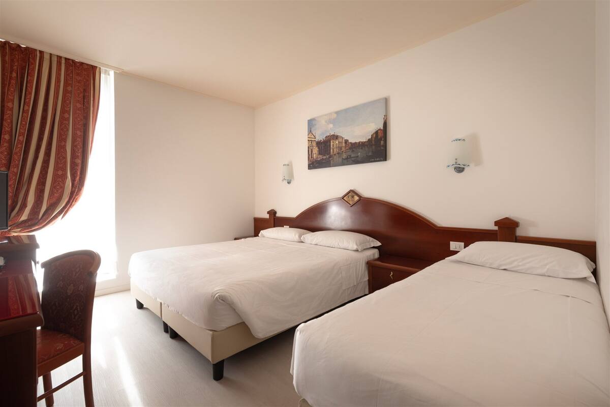 A hotel room with two single beds, a wooden headboard, a desk with a chair, and a painting on the wall.