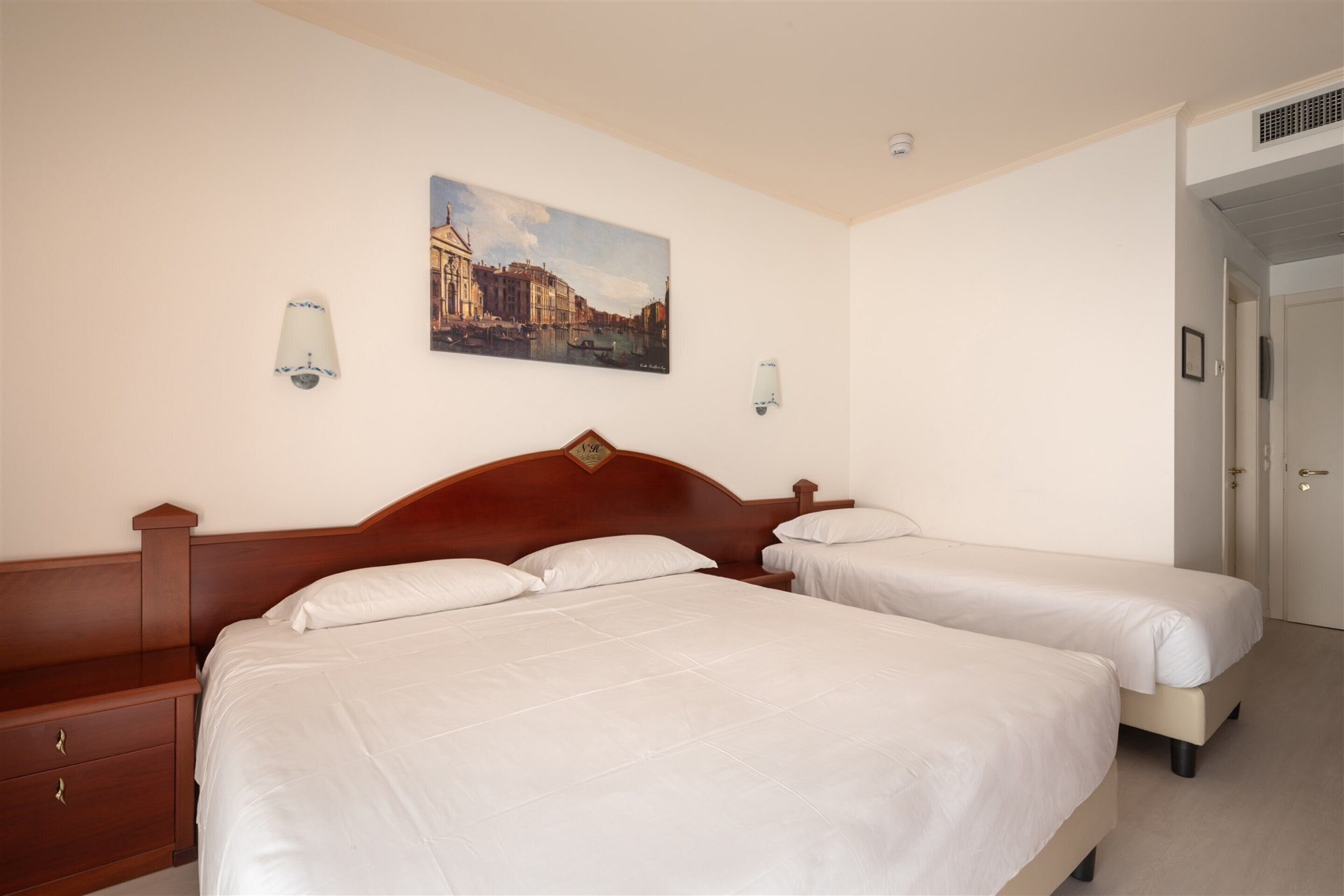 A hotel room with two beds, a wooden headboard, a painting of a cityscape, and a door to the right.