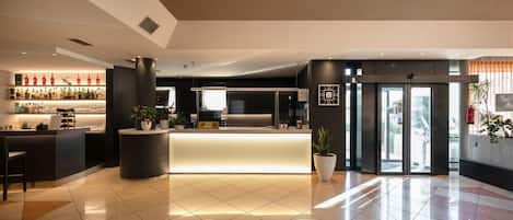 A modern hotel lobby with a reception desk, a bar area with various bottles, and a seating area with a potted plant.