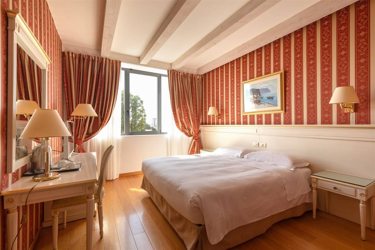 A hotel room with a bed, a desk with a lamp, a chair, a nightstand, and a window with curtains.