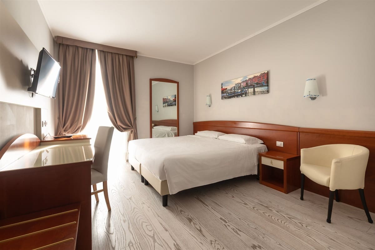 A hotel room with a bed, a desk, a chair, a TV, and a painting on the wall.