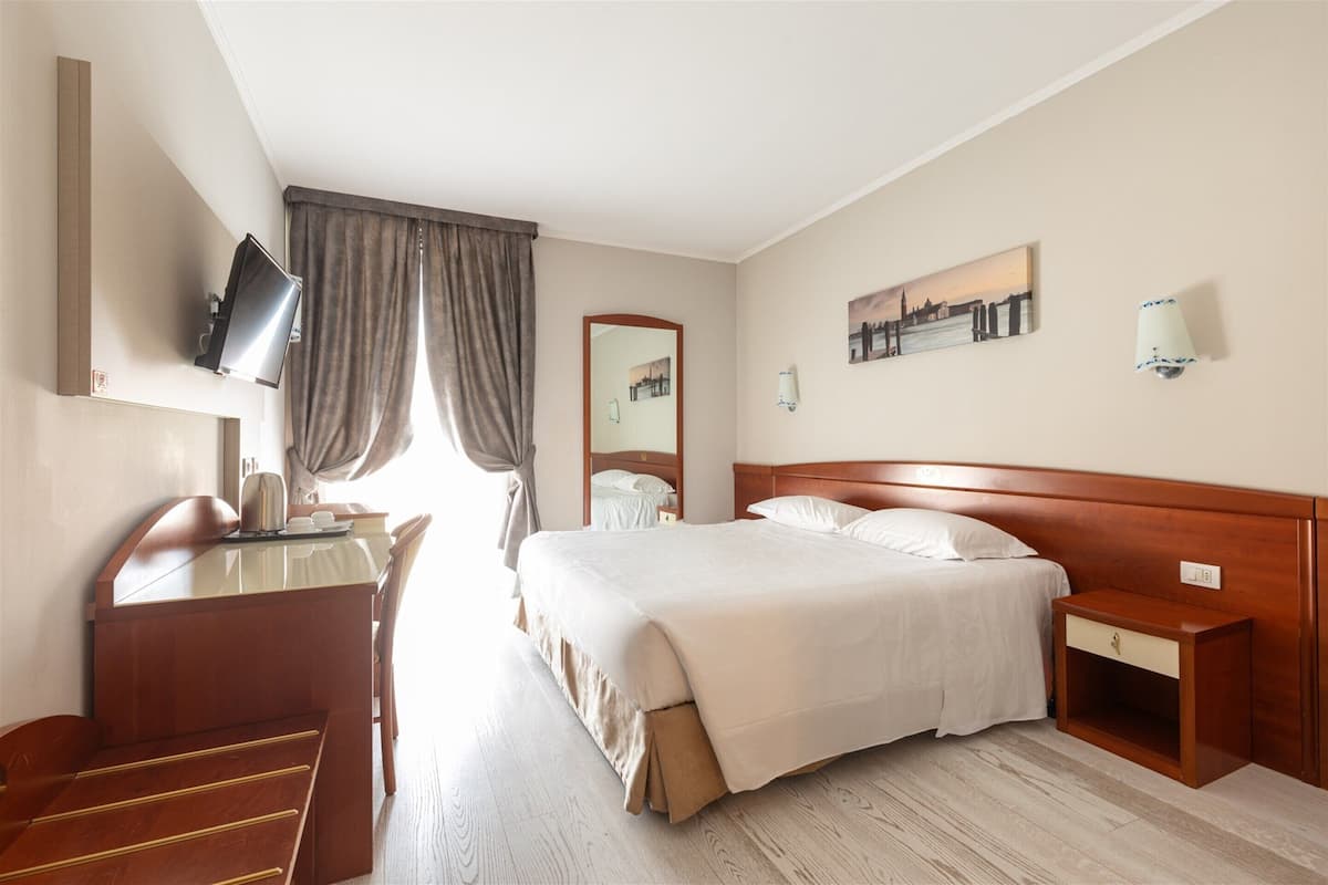 A hotel room with a bed, bedside table, TV, desk, chair, and a window with curtains.