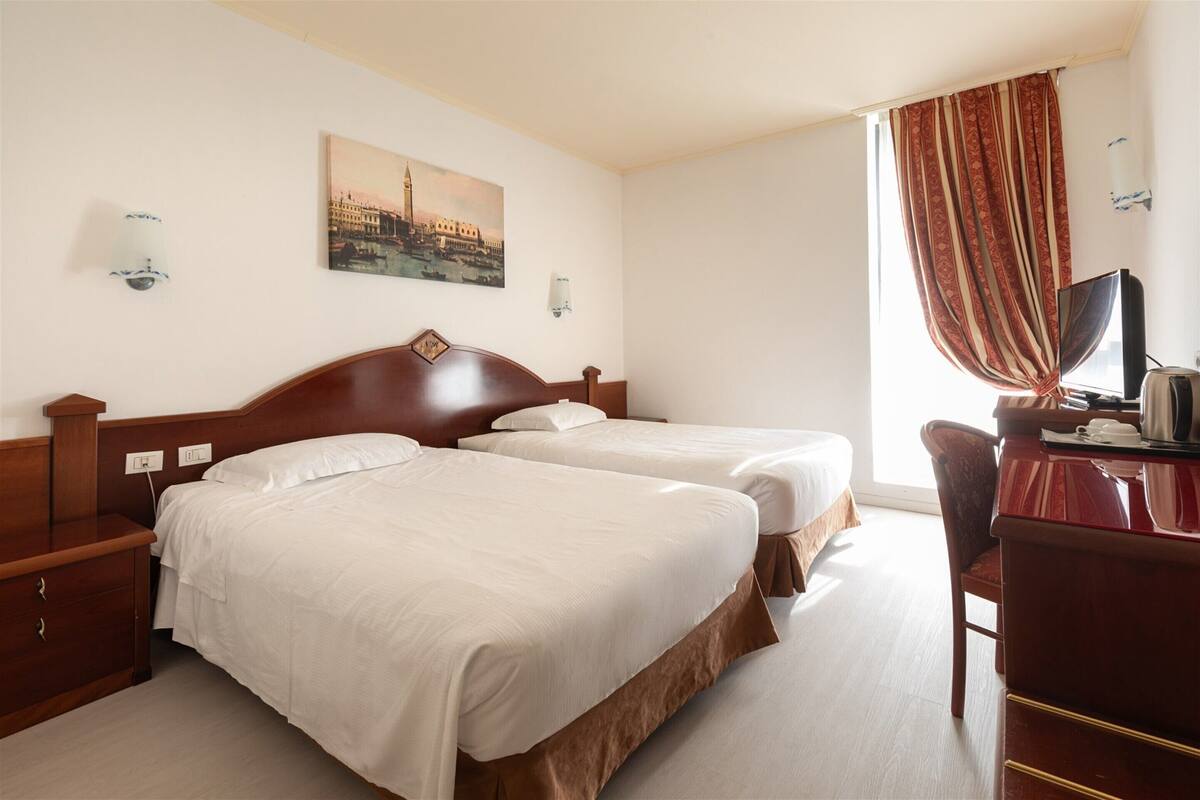 A hotel room with two beds, a desk, a chair, a TV, and a painting of a cityscape.