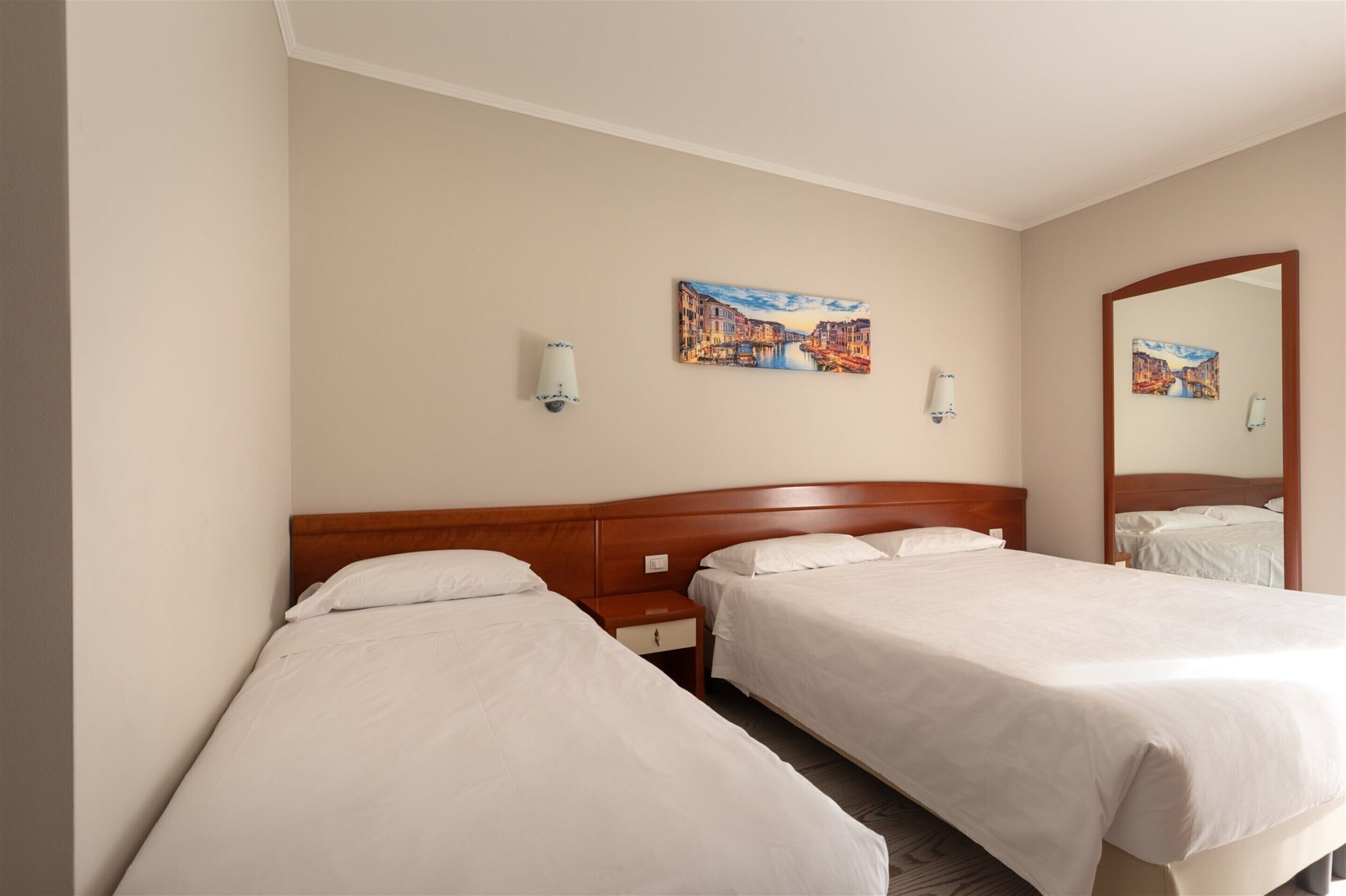 A hotel room with two single beds, a wooden headboard, a nightstand with a lamp, and two wall-mounted lamps.