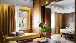 Una habitación de hotel moderna con un sofá, una mesita con un libro, una mesita de noche con un florero y una cama con un almohábil amarillo, además de una ventana con cortinas.