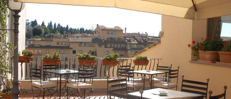 An outdoor terrace with tables and chairs, potted plants, and a view of buildings in the background.