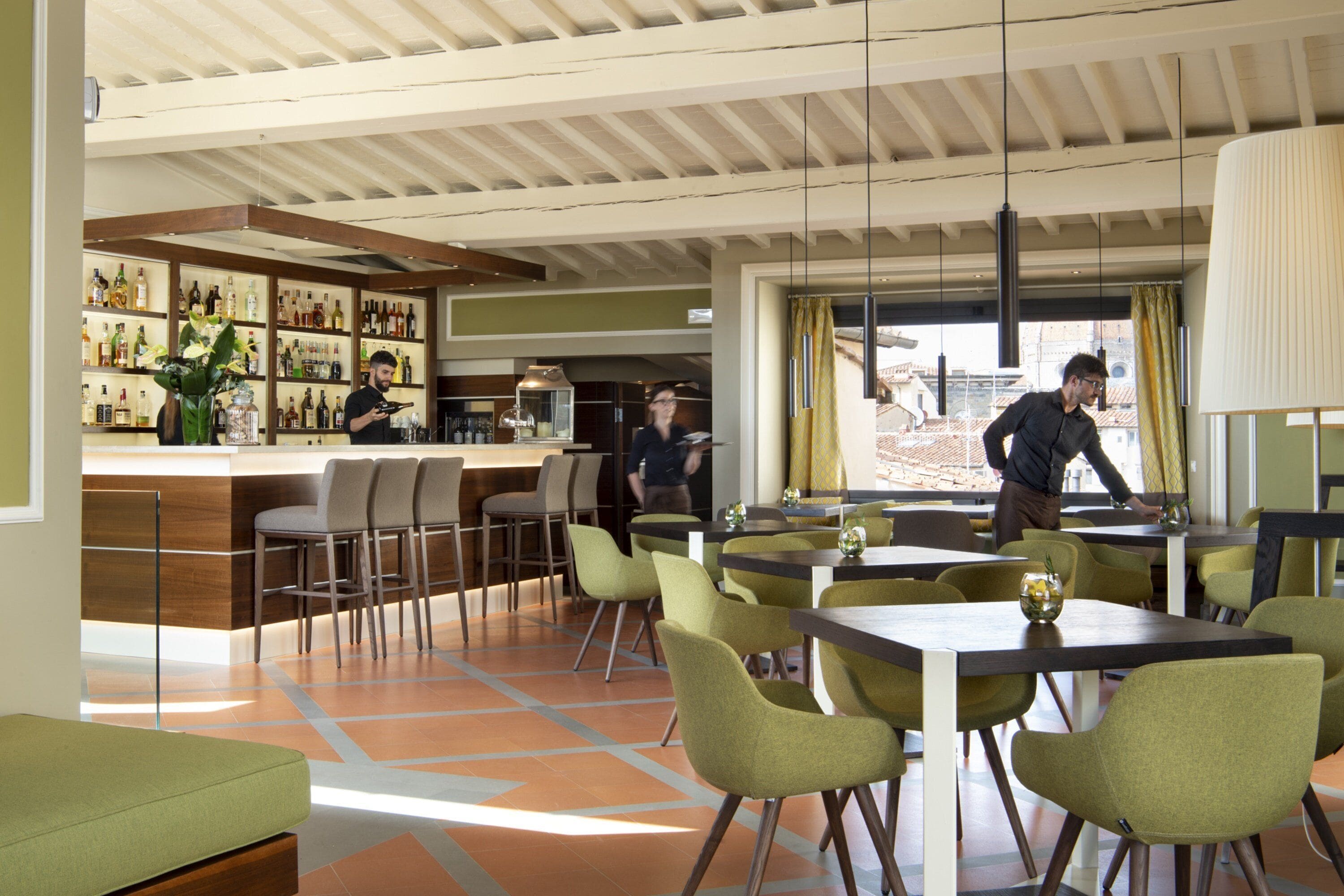 A modern dining area with green chairs and tables, a bar with shelves of bottles, and a view of buildings through large windows.