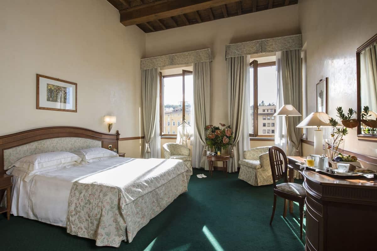 A hotel room with a large bed, two bedside tables, a desk with a chair, a window with curtains, and a view of buildings outside.
