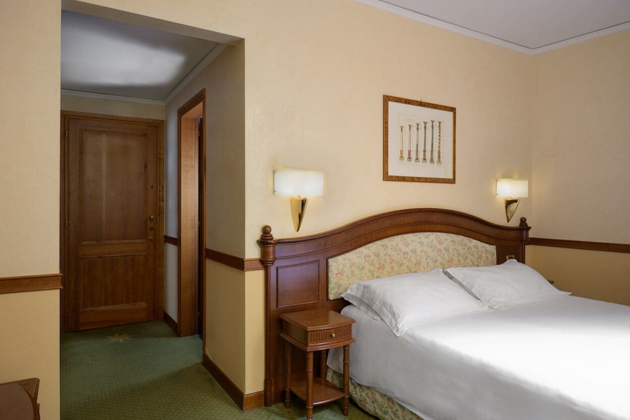 Habitación de hotel con puerta de madera, cama con sábanas blancas, mesita de noche con lámpara y un cuadro en la pared.