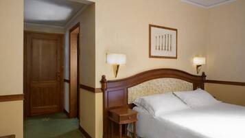 Habitación de hotel con puerta de madera, cama con sábanas blancas, mesita de noche con lámpara y un cuadro en la pared.