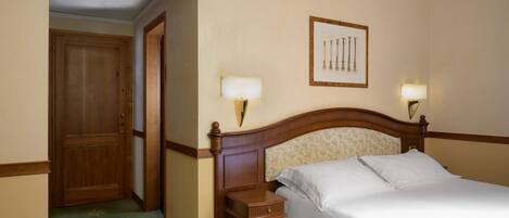Habitación de hotel con puerta de madera, cama con sábanas blancas, mesita de noche con lámpara y un cuadro en la pared.