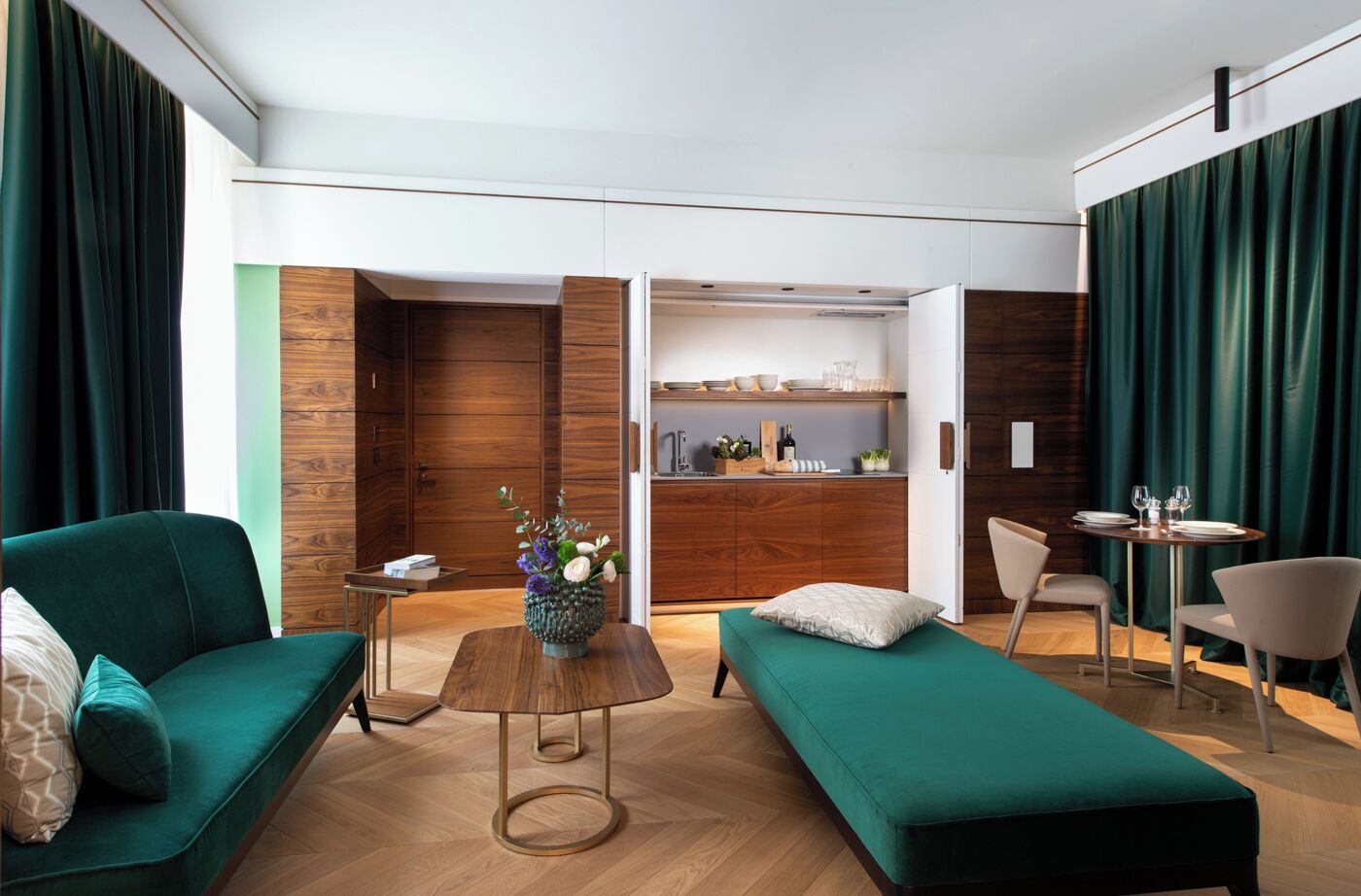Una habitación de hotel moderna con un sofá verde, una mesita redonda con un florero, un banco verde y una zona de comedor con una mesa y sillas.