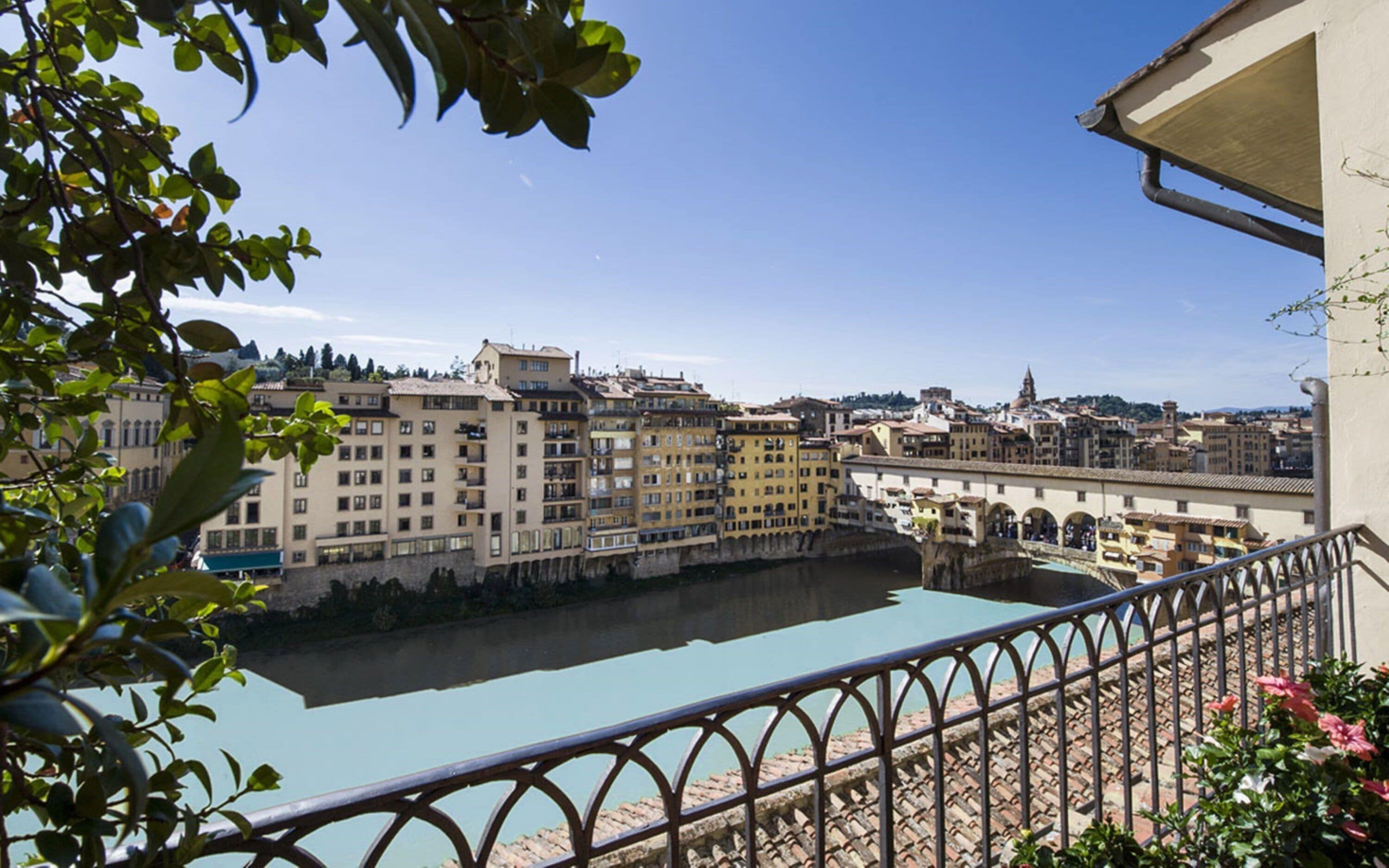 A view of a river with buildings on the opposite bank, a bridge, and a balcony with a railing.