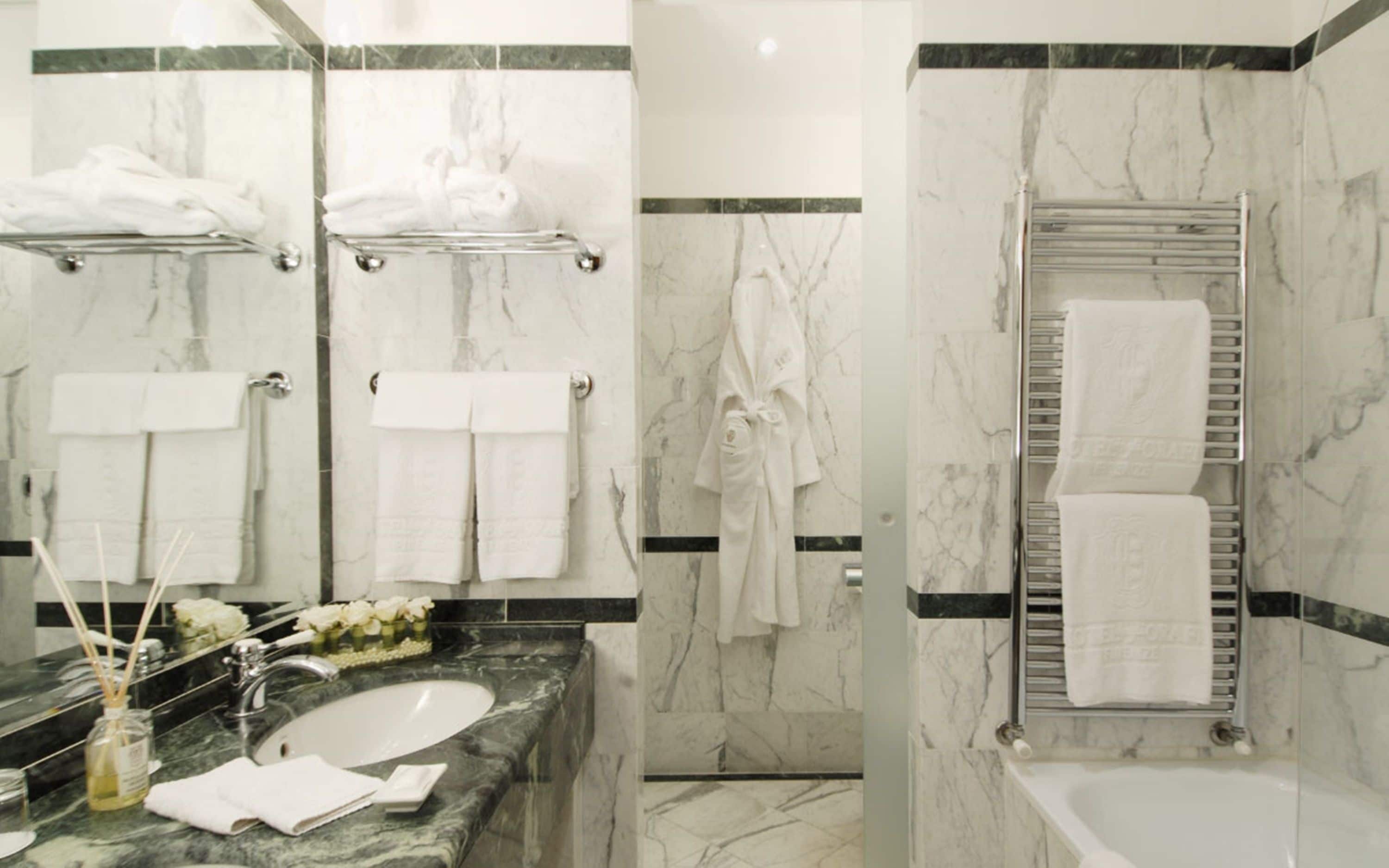 Un baño moderno con paredes de mármol, un lavamanos y una bañera. Hay varias toallas blancas y una bata colgadas en la pared.