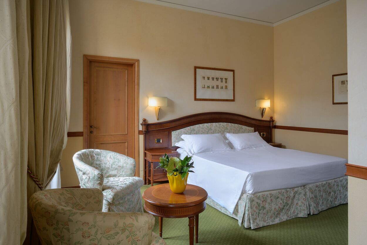 A hotel room with a bed, two chairs, a wooden nightstand, a lamp, and a framed picture on the wall.