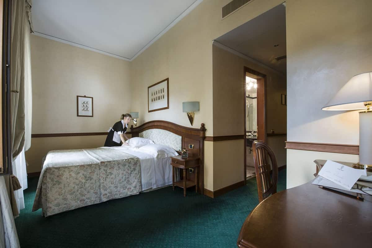 A hotel room with a bed, a desk, a chair, a nightstand, a lamp, and a mirror.