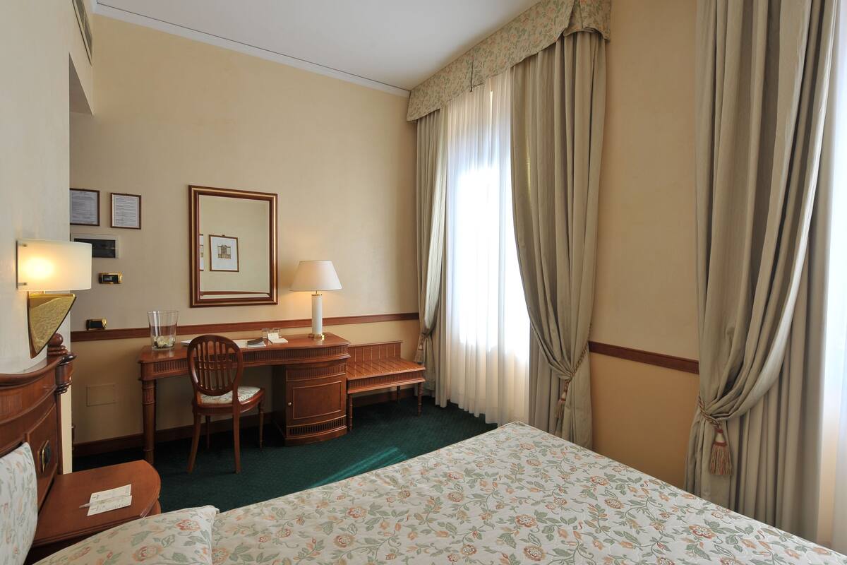 A hotel room with a bed, a desk, a chair, a lamp, and a mirror.