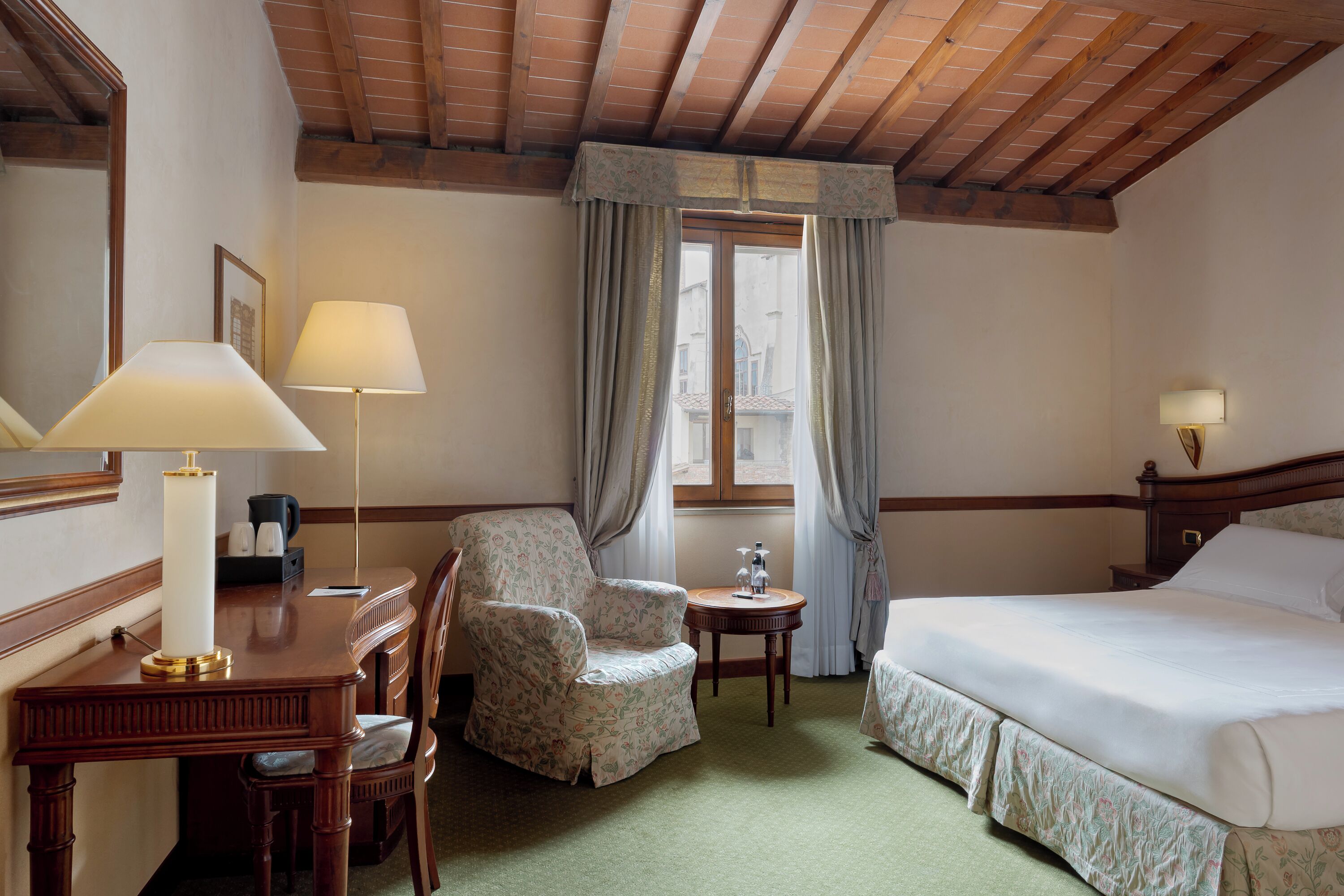 A hotel room with a bed, a desk with a lamp, a chair, a small table, and a window with curtains.