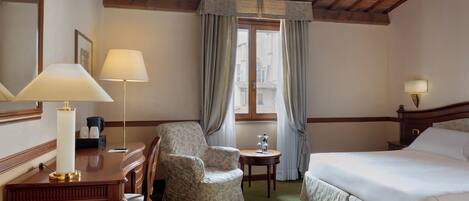 Une chambre d’hôtel comprenant un lit, un bureau avec une lampe, une chaise, une petite table et une fenêtre avec des rideaux.