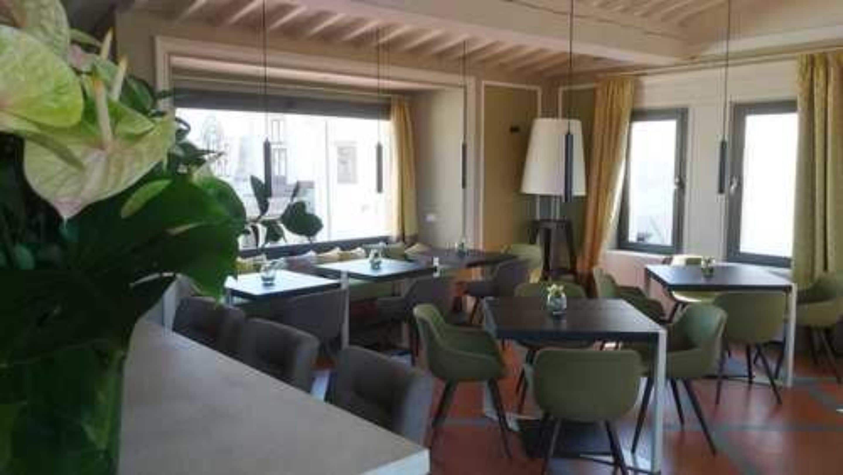 A hotel dining area with multiple tables and chairs, large windows with curtains, and a ceiling with wooden beams.
