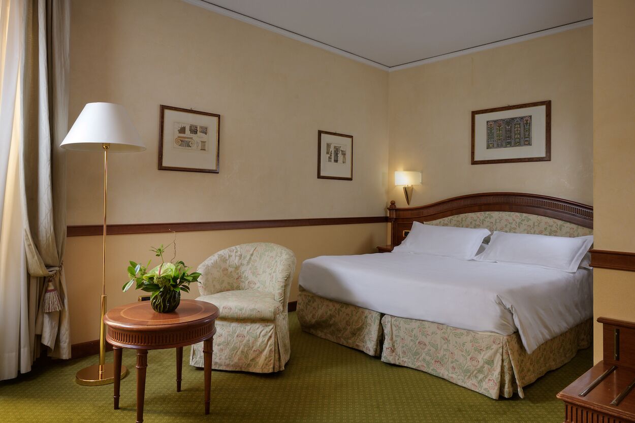 Quarto de hotel com uma cama, uma cadeira, uma mesa com uma planta e duas molduras na parede.