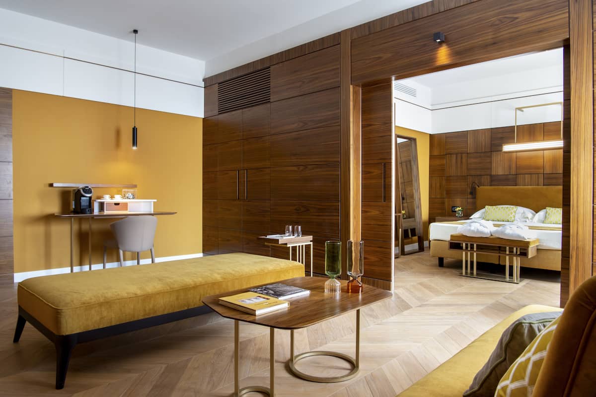 A modern hotel room with a wooden accent wall, a bed, a bench, a small table with a book, and a mirror.
