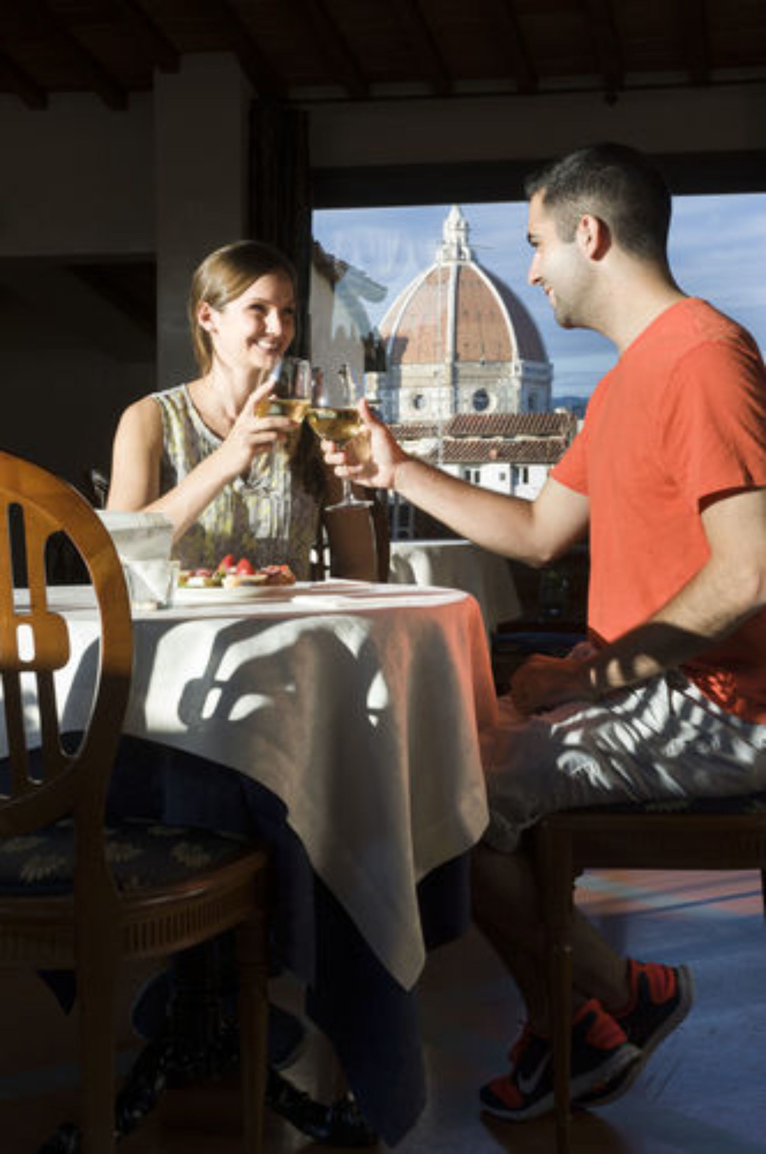 A dining table with a white tablecloth, two people toasting with wine glasses, and a view of a building with a dome in the background.