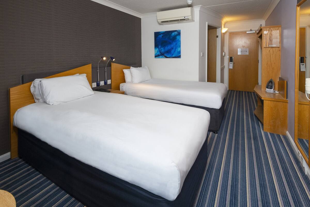 A hotel room with two beds, a blue-striped carpet, a wooden headboard, and a wall-mounted air conditioner.